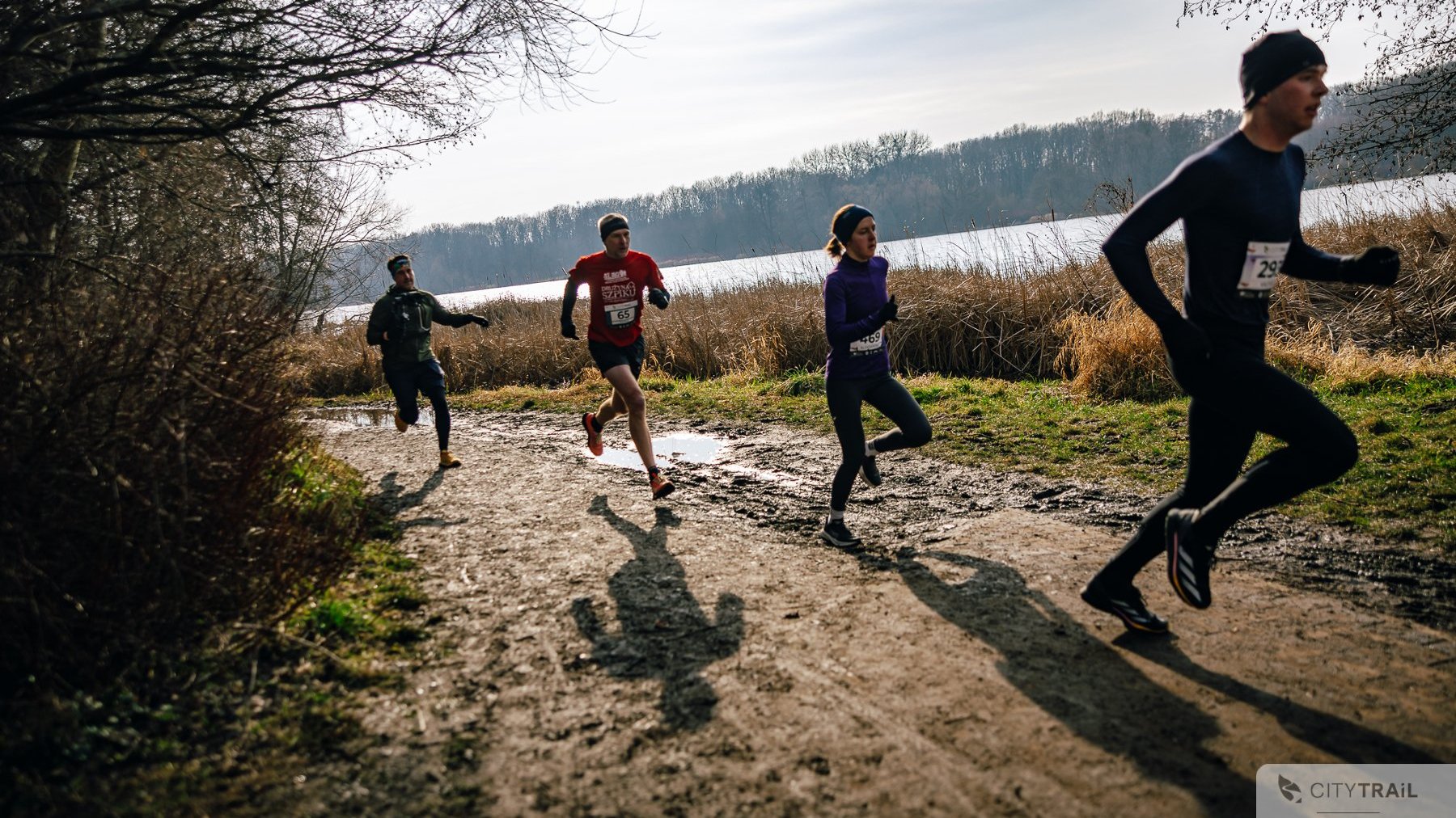 Bieg City Trail, fot. Andrzej Olszanowski - 3 zawodników i zawodniczka biegną w słonecznej aurze po grząskim terenie. W tle las oraz jezioro