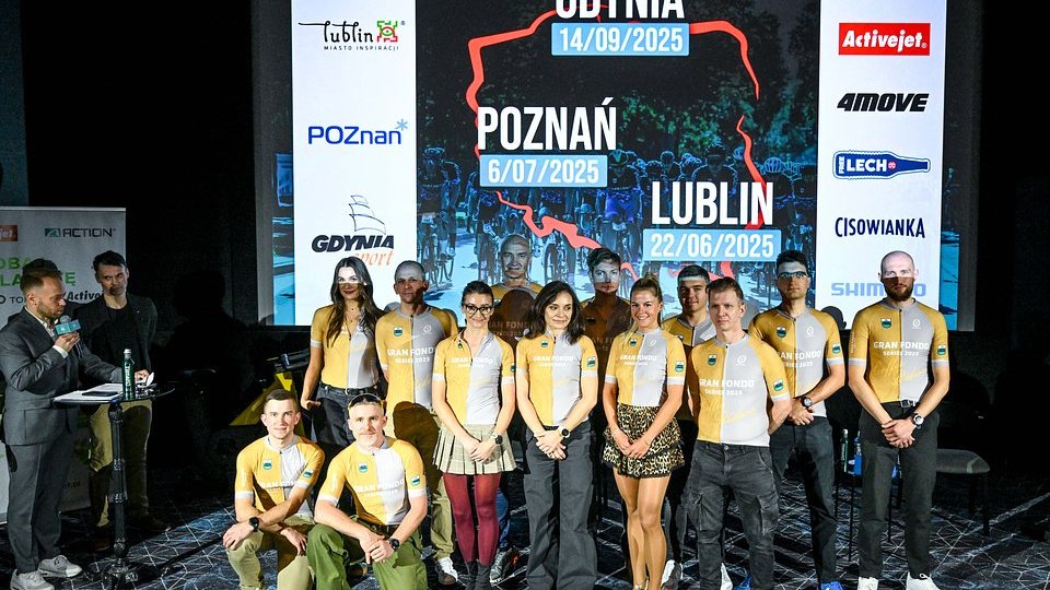 Konferencja prasowa przed cyklem Gran Fondo, fot. Barłomiej Zborowski - na pierwszym planie animatorzy sportu kolarskiego w koszulkach, w których będą jeździć w Gran Fondo. W tle plansza ze sponsorami, terminami i lokalizacjami wyścifgu. Z lewej strony osoba prowadząca konferencję z mikrofonem stoi w garniturze