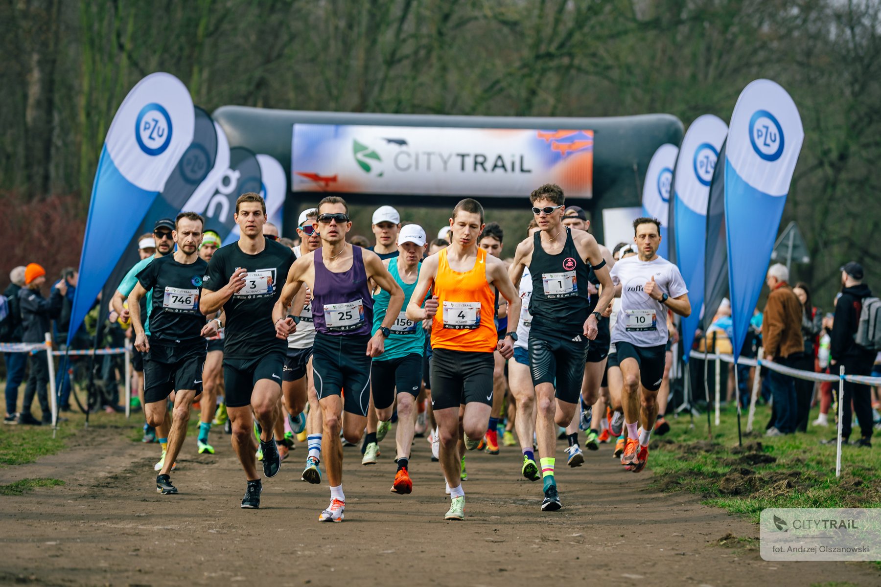 Ostatni bieg City Trail w Poznaniu 2024/2025, fot. Andrzej Olszanowski - biegacze na starcie biegu, w tle widać bramę zawodów oraz las, a po bokach reklamy sponsorów. Z boku widać obserwujących start kibiców. - grafika artykułu