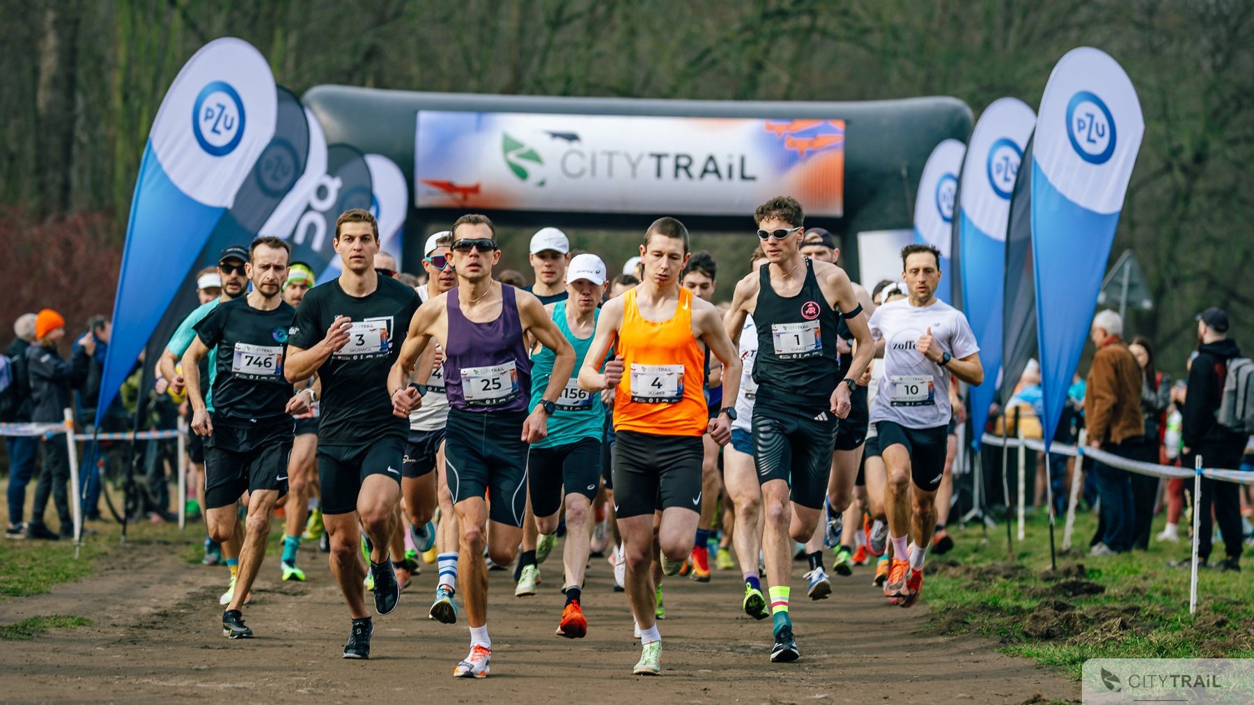 Ostatni bieg City Trail w Poznaniu 2024/2025, fot. Andrzej Olszanowski - biegacze na starcie biegu, w tle widać bramę zawodów oraz las, a po bokach reklamy sponsorów. Z boku widać obserwujących start kibiców.