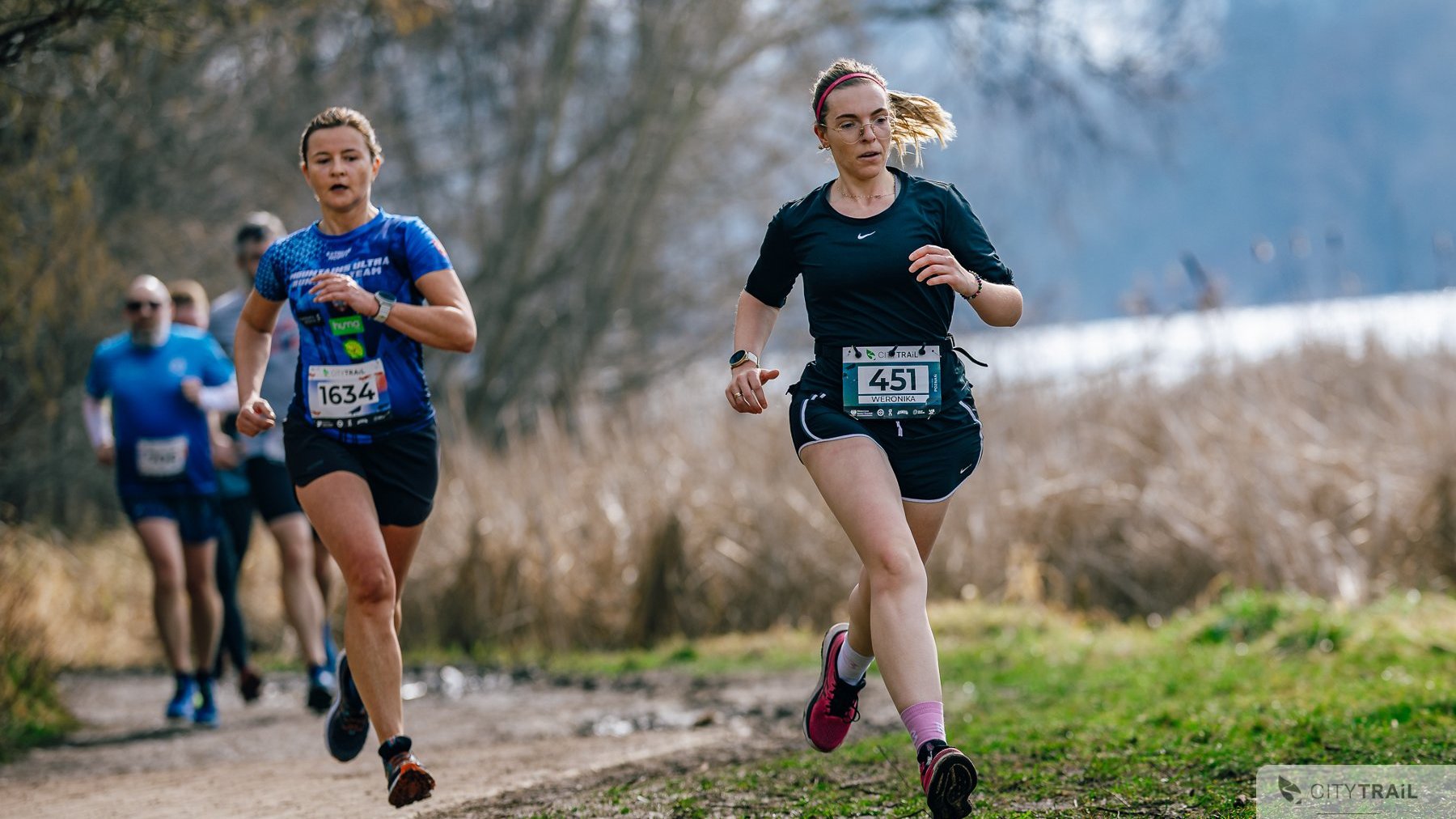 Ostatni bieg City Trail w Poznaniu 2024/2025, fot. Andrzej Olszanowski - na pierwszym planie 2 biegnące kobiety, jedna w czarnym, a druga w niebieskim stroju. W tle widać jezioro i las w słonecznej aurze