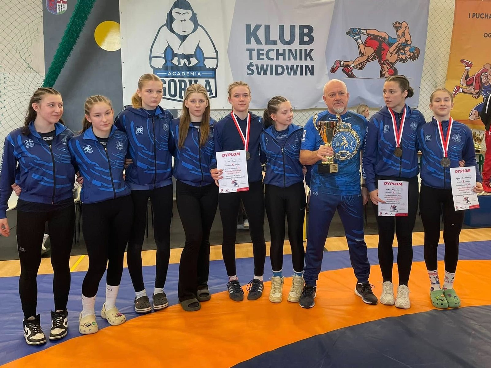 Zawodniczki Sobieskiego Poznań z medalami Pucharu Polski Kadetek w zapasach, fot. Maciej Kula - w strojach klubowych stoją zawodniczki Sobieskiego Poznań, 3 z nich mają medale i dyplomy. W środku stoi trener, który trzyma puchar. - grafika artykułu