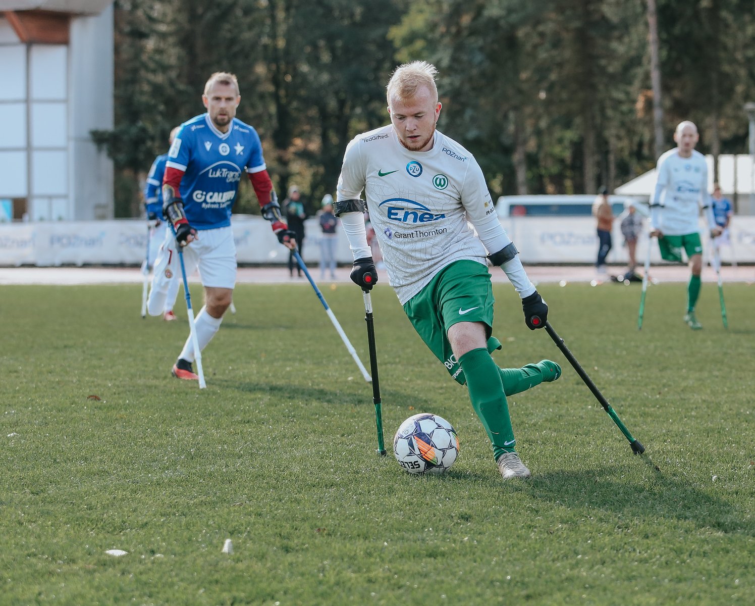 Zawodnicy sekcji amp futbolu Warty Poznań, fot. Tomasz Stefanik/Amp Futbol Polska - na pierwszym planie zawodnik sekcji amp futbolu Warty Poznań w białej koszulce i zielonych spodenkach przy piłce. W tle na boisku inny zawodnik drużyny Warty Poznań oraz zawodnicy przeciwnej drużyny w niebieskich koszulkach i białych spodenkach. W tle widać także drzewa. - grafika artykułu