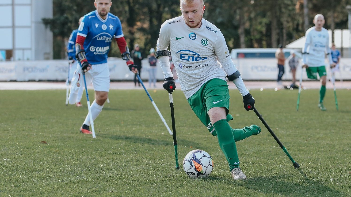 Zawodnicy sekcji amp futbolu Warty Poznań, fot. Tomasz Stefanik/Amp Futbol Polska - na pierwszym planie zawodnik sekcji amp futbolu Warty Poznań w białej koszulce i zielonych spodenkach przy piłce. W tle na boisku inny zawodnik drużyny Warty Poznań oraz zawodnicy przeciwnej drużyny w niebieskich koszulkach i białych spodenkach. W tle widać także drzewa.