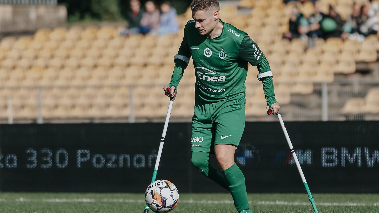 Zawodnicy sekcji amp futbolu Warty Poznań, fot. Tomasz Stefanik/Amp Futbol Polska - przy piłce zawodnik sekcji amp futbolu Warty Poznań w zielonej koszulce i zielonych spodenkach. W tle trybuny z żółtymi krzesełkami i nielicznymi kibicami siedzącymi na nich