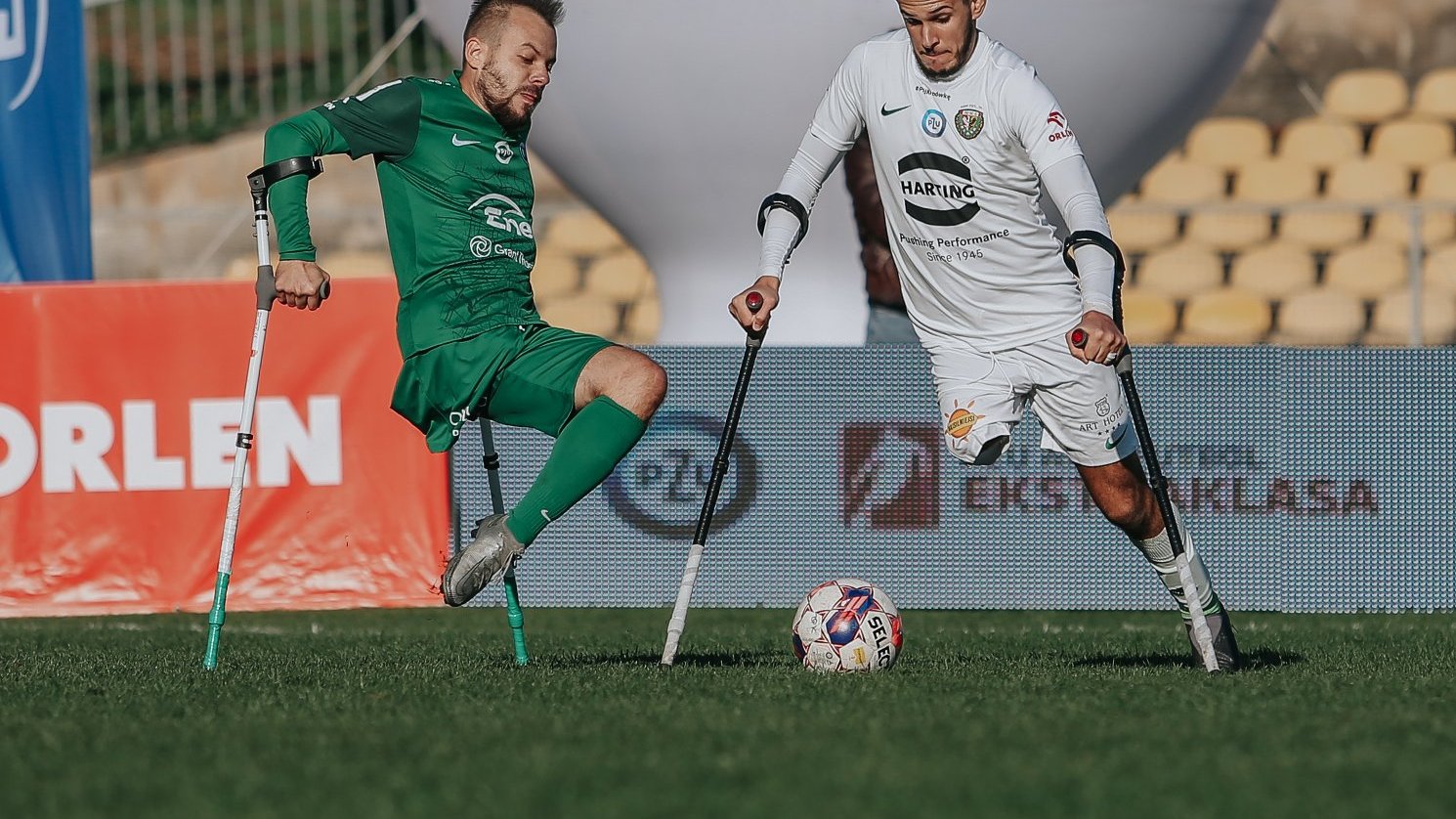 Zawodnicy sekcji amp futbolu Warty Poznań, fot. Tomasz Stefanik/Amp Futbol Polska - o piłkę walczy zawodnik amp tubolu Warty Poznań w zielonej koszulce i zielonych spodenkach oraz zawodnik drużyny w przeciwnej w białej koszulce i białych spodenkach. W tle widać biały balon pneumatycznych oraz puste krzesełka na trybunach w kolorze żółtym