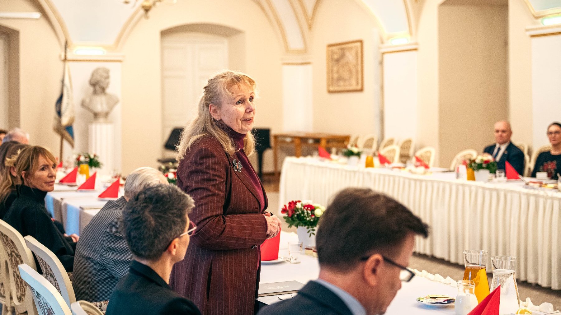 Formalne spotkanie w eleganckim pomieszczeniu z łukowymi sufitami i żyrondami. Wokół stołu pokrytego białym fartuchem siedzą osoby, a na stole znajdują się czerwone ręczniki, kwiaty i dzbanki z sokiem lub wodą. Jedna osoba stoi i wydaje się prowadzić prezentację lub mówić do grupy. W pokoju są także popiersie i ujęte w ramy dzieła sztuki na ścianach.