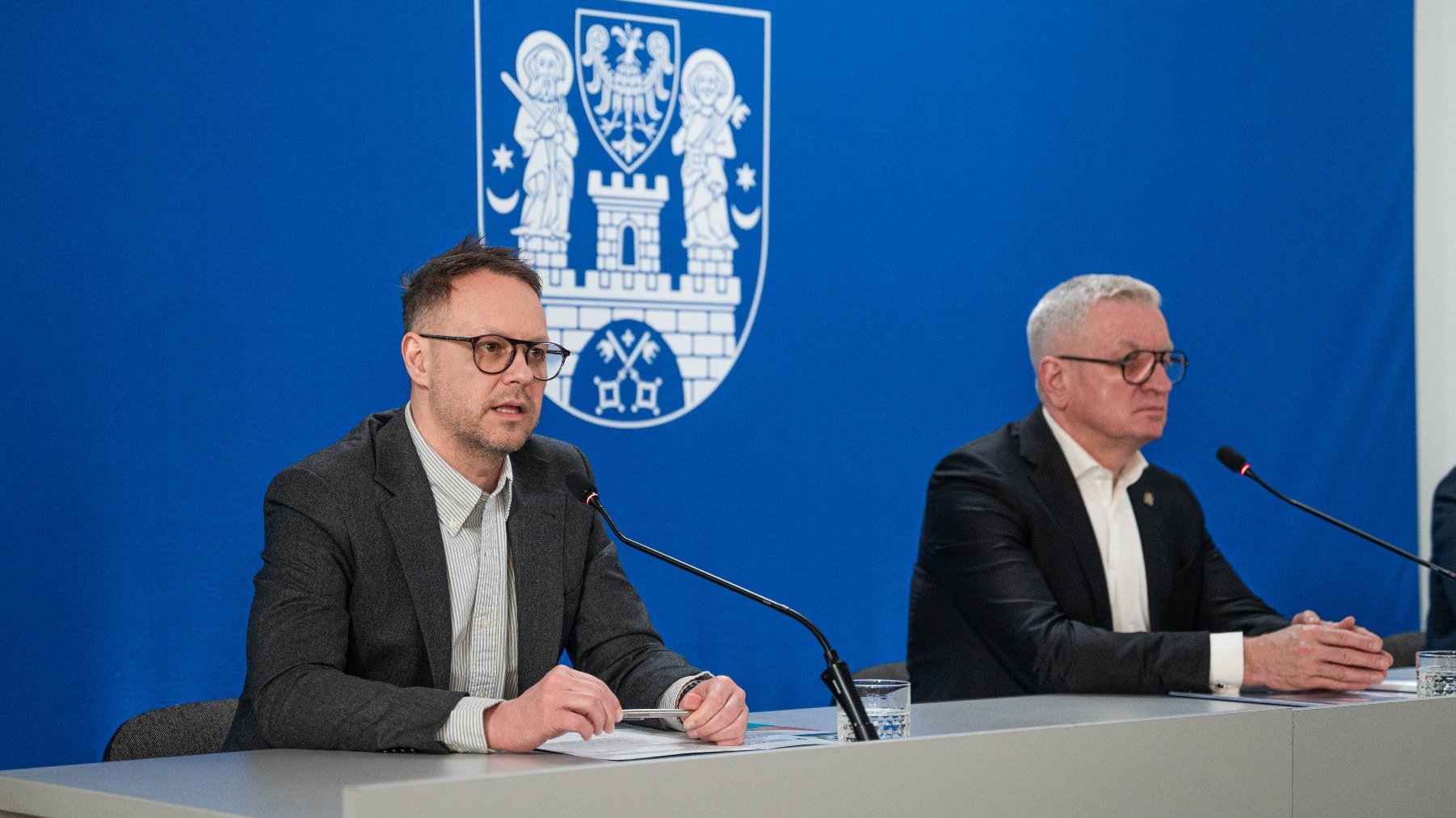 Na zdjęciu dwóch mężczyzn za stołem konferencyjnym, jeden z nich mówi coś do mikrofonu