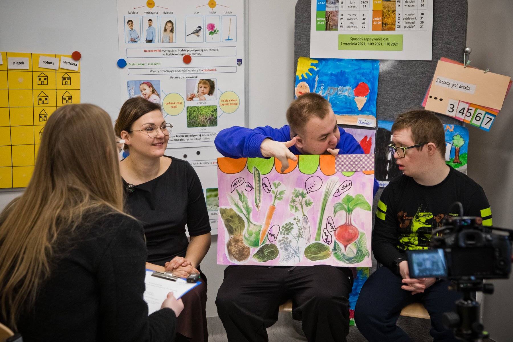 Zdjęcie grupy osób w przestrzeni edukacyjnej. Na ścianach widoczne różne materiały edukacyjne i plakaty, w tym kalendarz z dniami tygodnia po polsku ("piątek", "sobota", "niedziela") oraz ilustracje przedstawiające warzywa (m.in. marchewkę, rzodkiewkę) z ich nazwami. Jedna z osób trzyma duży, kolorowy plakat z rysunkami warzyw. - grafika artykułu