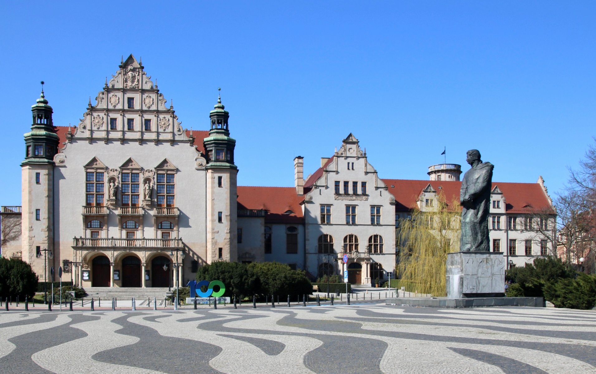 Zdjęcie przedstawia budynek rektoratu Uniwersytetu im. Adama Mickiewicza w Poznaniu na tle niebieskiego nieba. Przed budynkiem widać brukowany plac oraz pomnik Adama Mickiewicza. - grafika artykułu