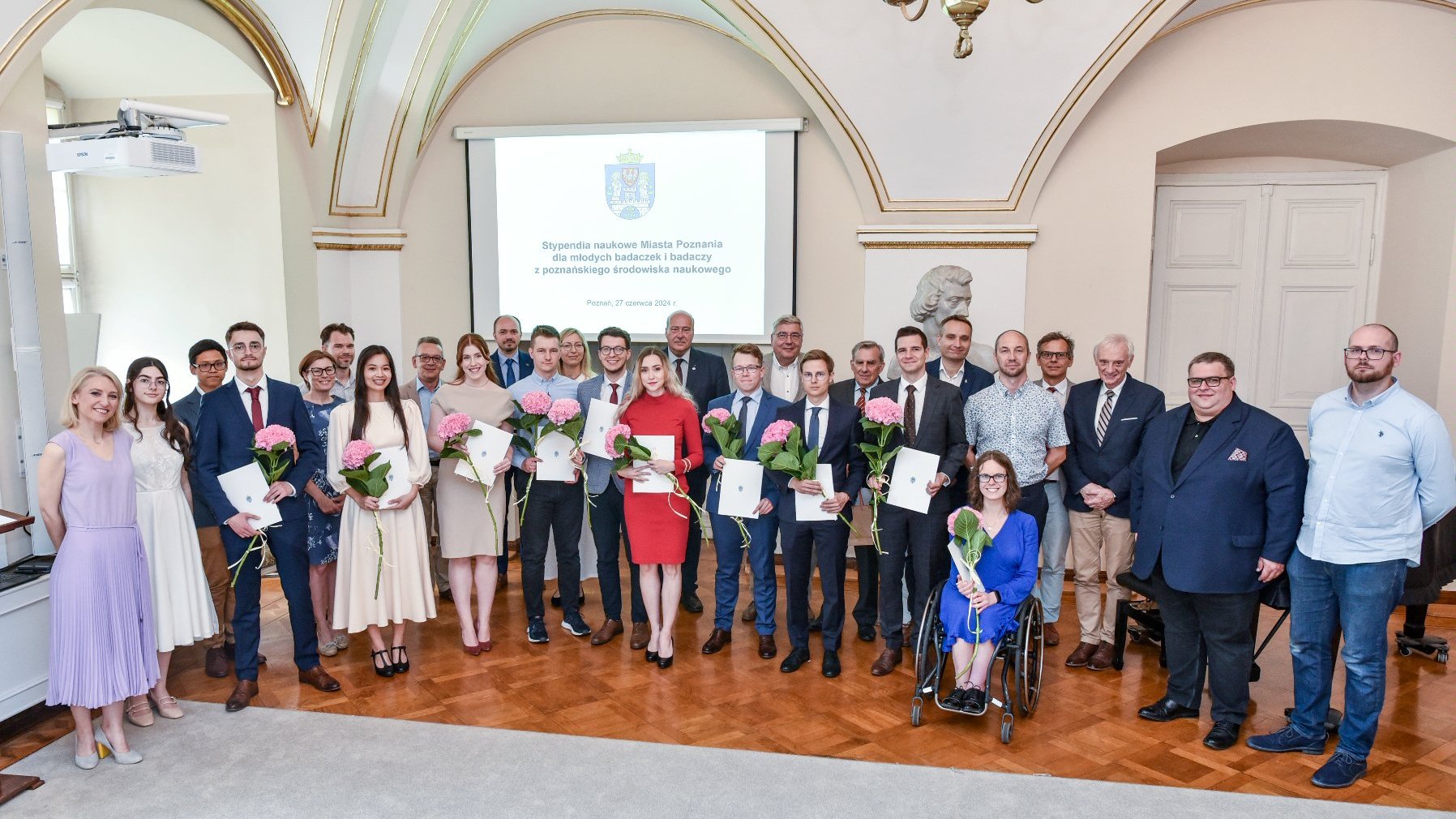 zdjęcie przedstawia grupę elegancko ubranych osób w sali urzędu miasta. W pierwszym rzędzie znajdują się laureatki i laureaci stypendium, którzy trzymają w rękach dyplomy i kwiaty.