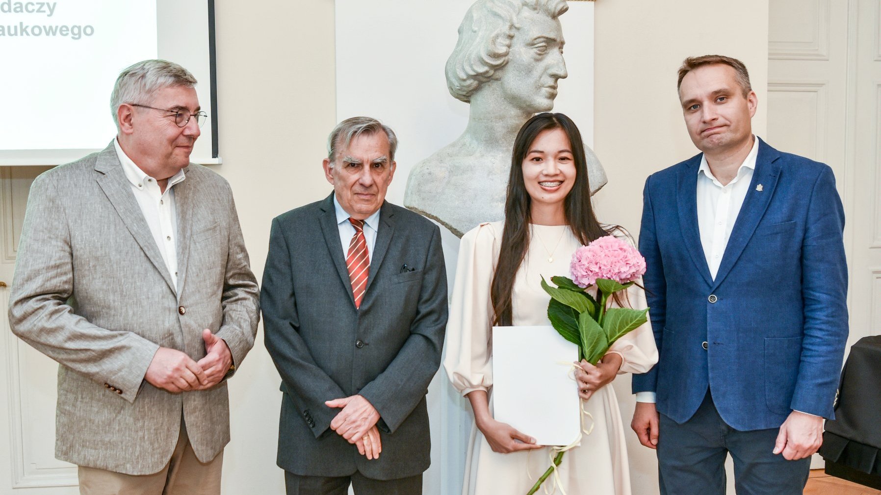 zdjęcie przedstawia cztery stojące osoby, w tym trzech mężczyzn i jedną kobietę, laureatkę stypendium, która trzyma przed sobą dyplom i kwiat