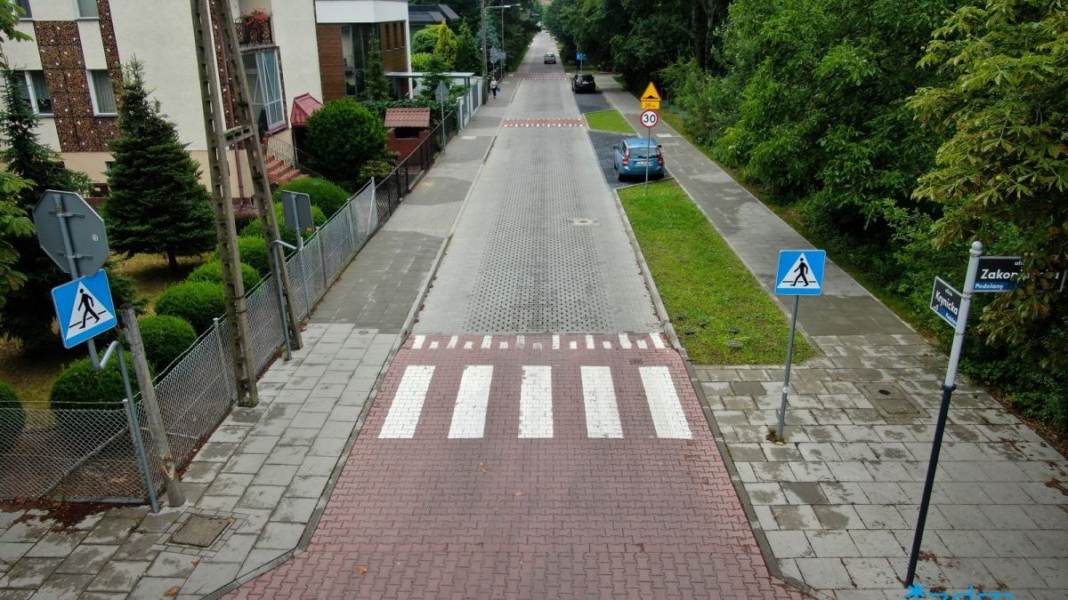 Zdjęcie przedstawia drogę lokalną. Po jednej jej stronie widać zabudowania, po drugiej drzewa.