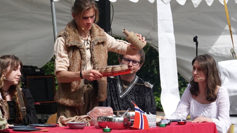 Zdjęcie przedstawia chłopaka w charakterystycznym stroju, m.in. w futrzanej kamizelce nalewającego coś z butelki do kubków. Obok siedzą osoby podobnie ubrane.