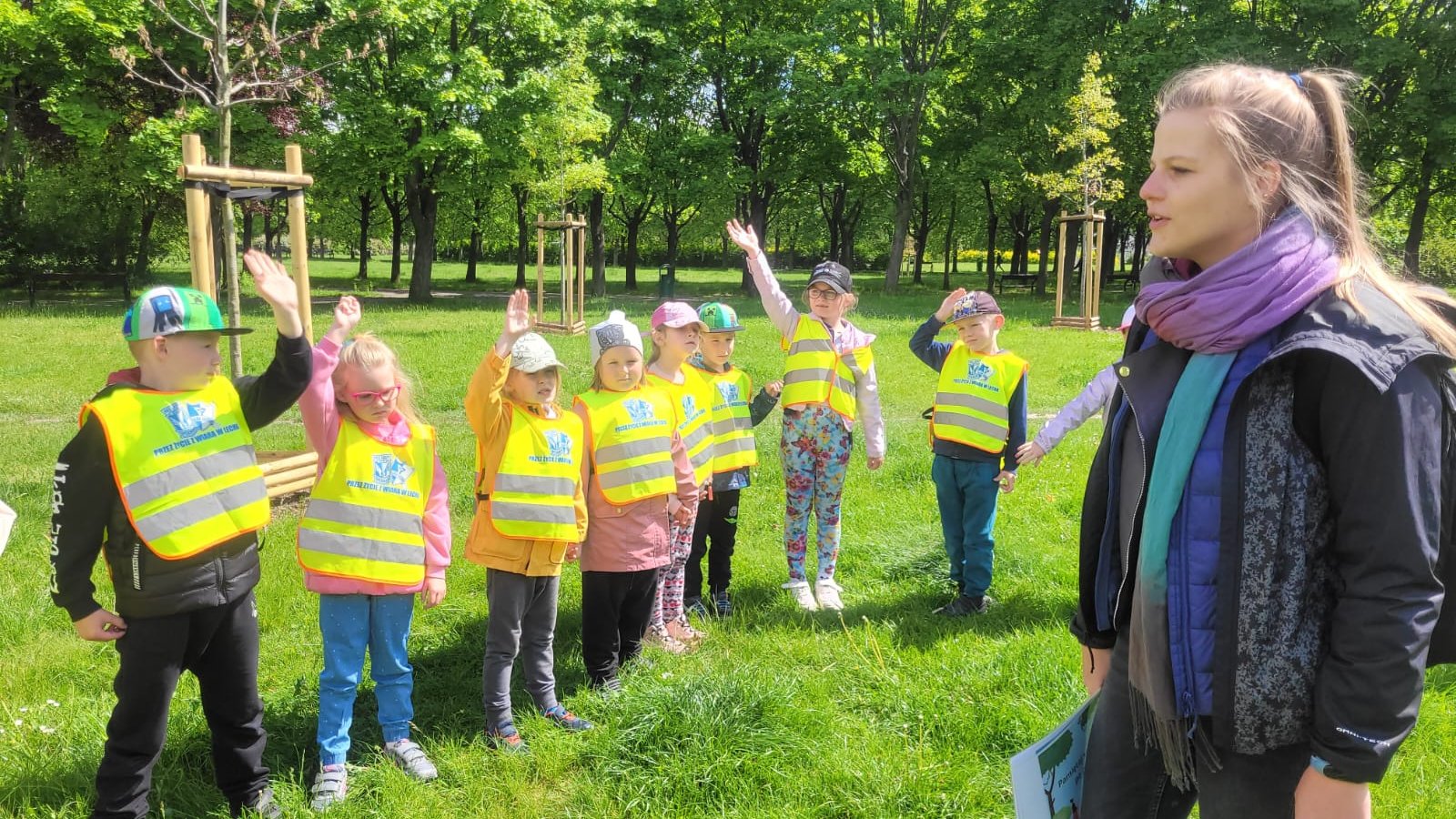 Zdjęcie przedstawia grupę dzieci i opiekunkę w parku.