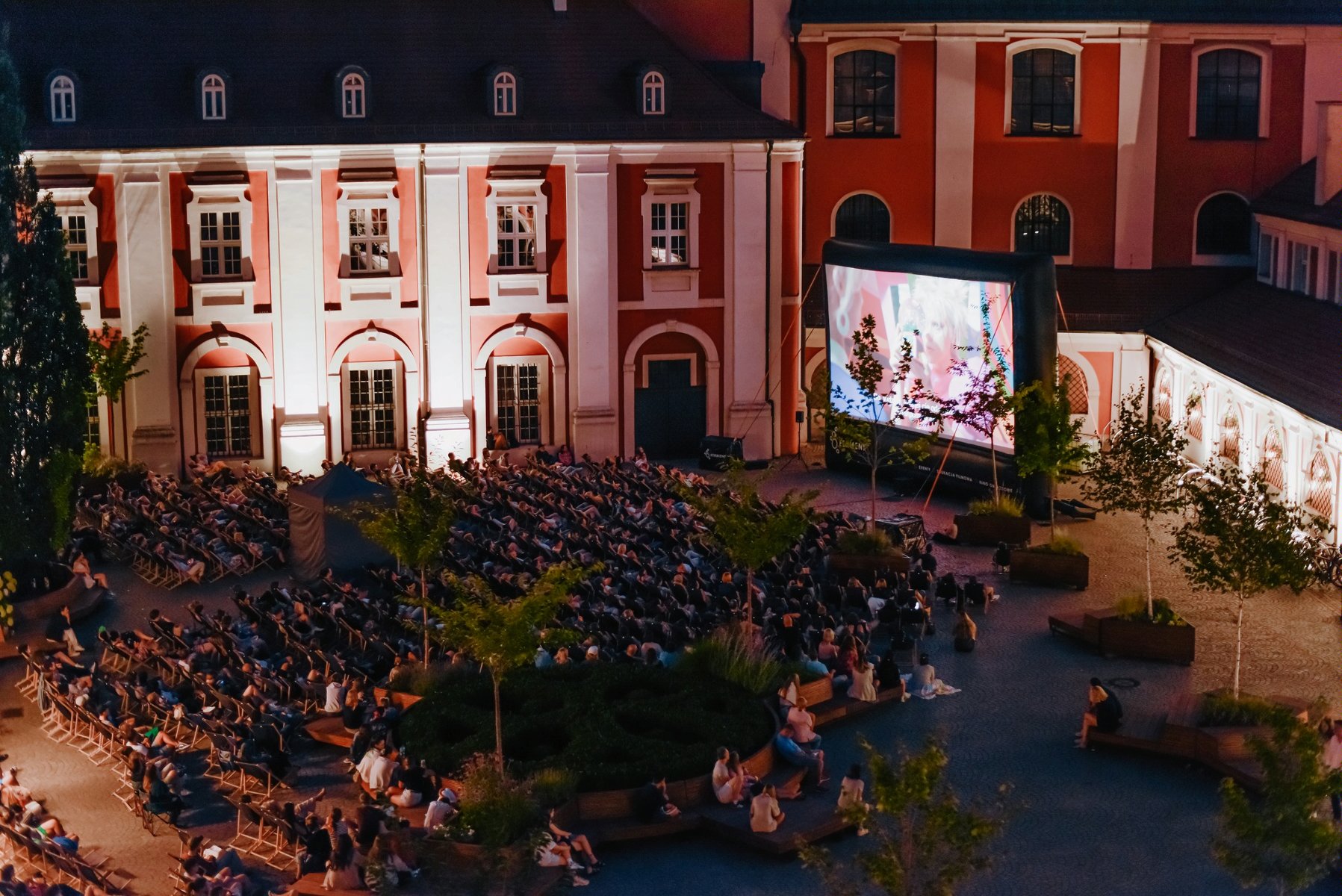 Zdjęcie przedstawia ludzi na leżakach oglądających film na telebimie na dziedzińcu urzędu miasta. - grafika artykułu