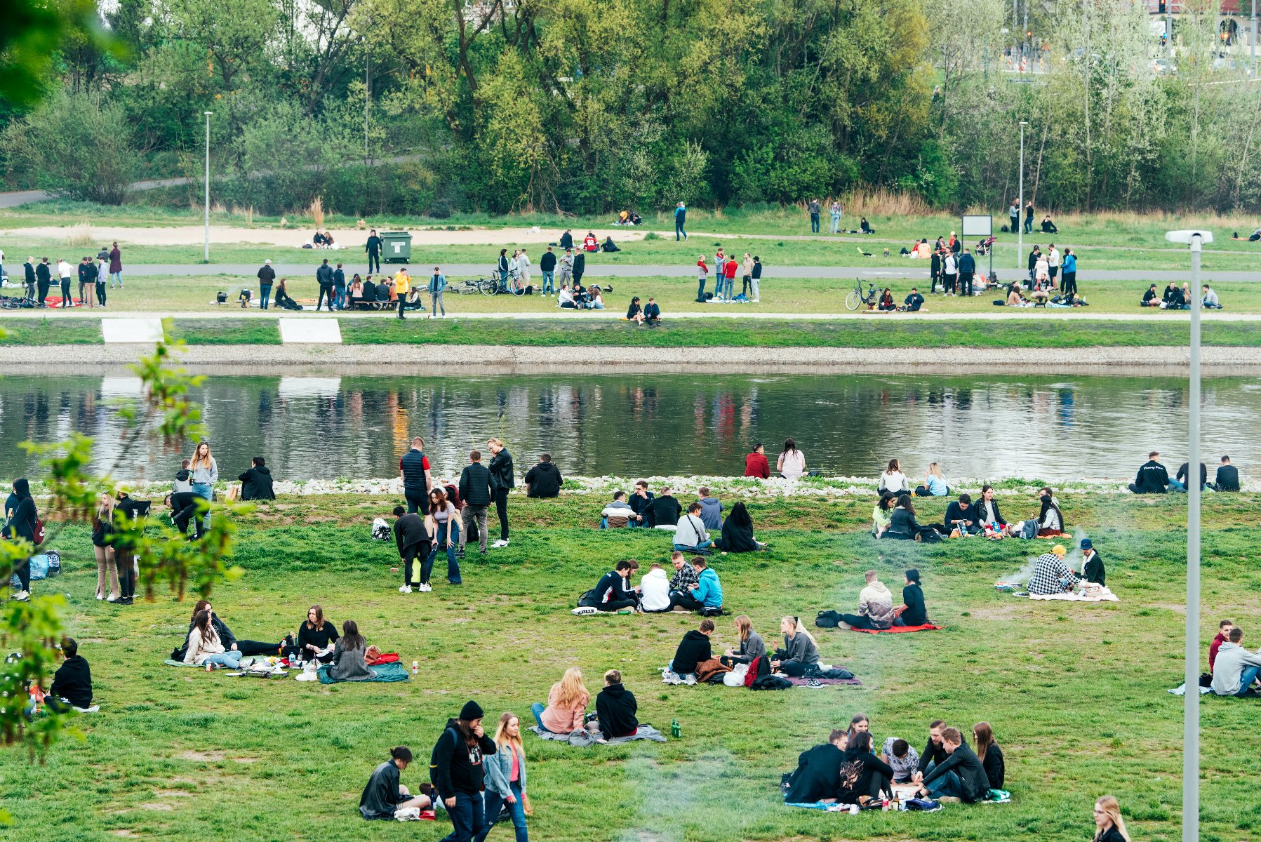 Zdjęcie przedstawia ludzi nad rzeką. Niektórzy siedzą na kocach, inni stoją i rozmawiają. - grafika artykułu