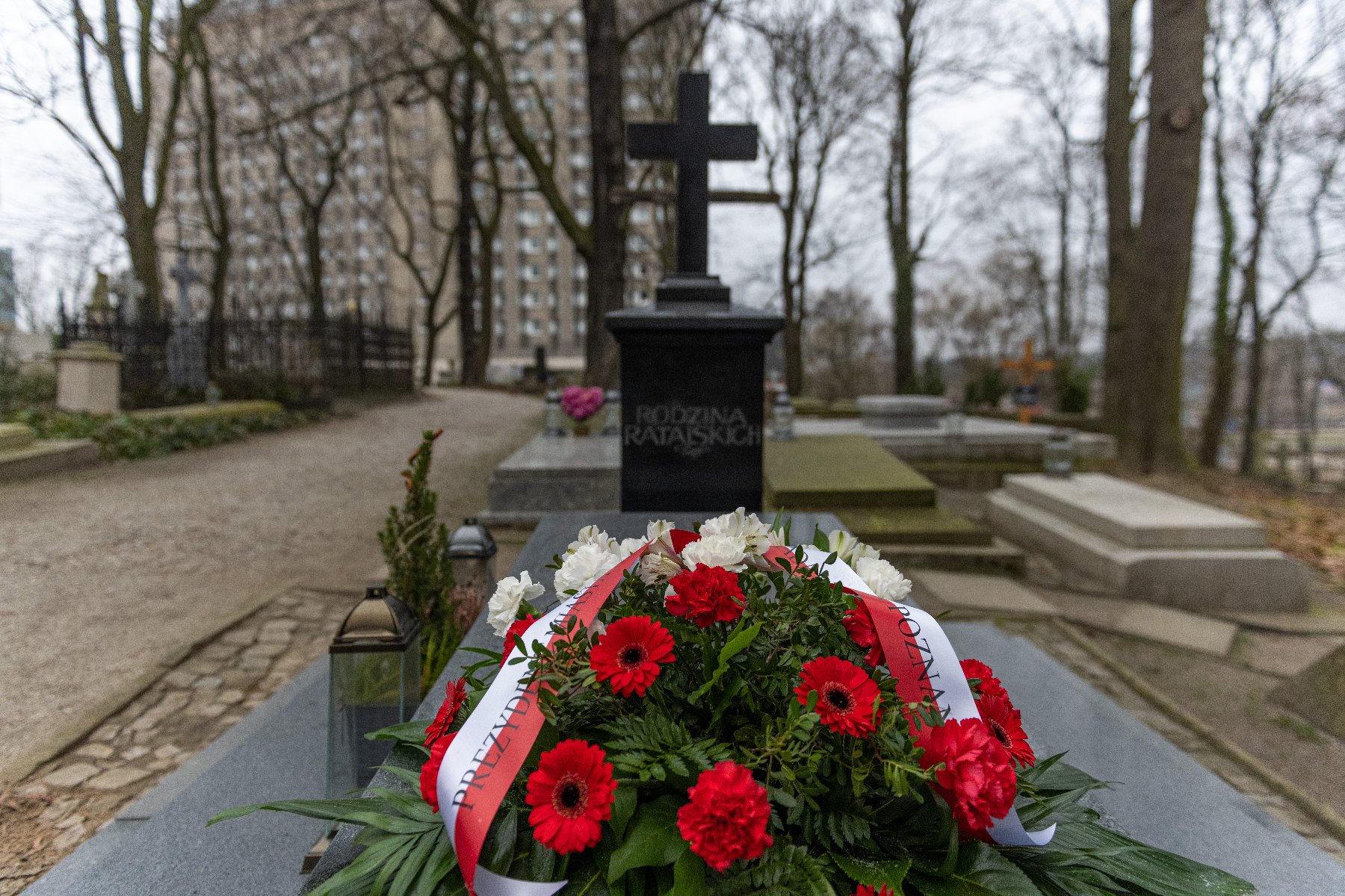 Cmentarz. Na środku grobowiec udekorowany kwiatami - czerwone i białe goździki i gerbery. Wstęga biała z czerwonymi napisami. Na czarnej płycie nagrobnej z krzyżem widnieje napis: "RODZINA RATAJSKICH". W tle inne groby, drzewa bez liści i wysoki budynek. Zima - brak liści na drzewach. - grafika artykułu