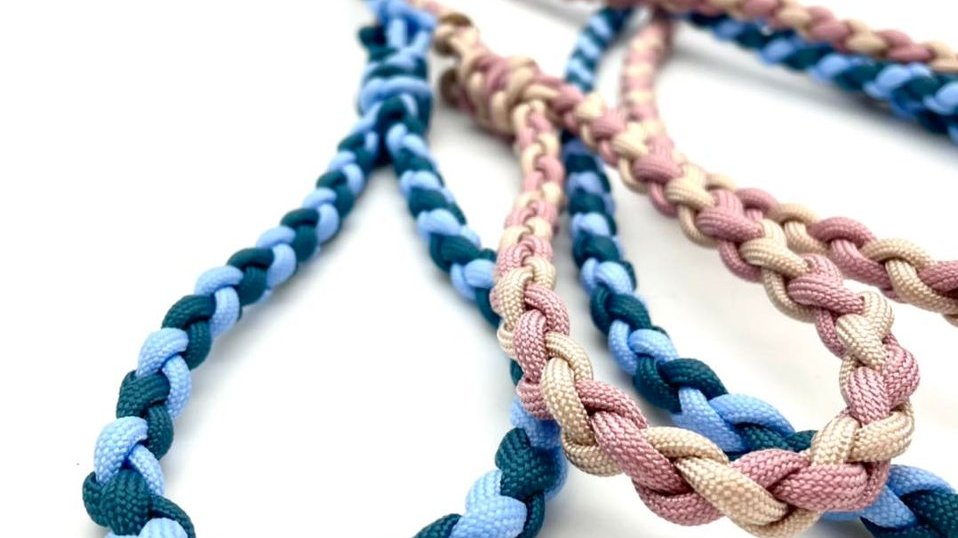 Dwie kolorowe smycze leżą na białym tle. Jedna jest skręcona z niebieskich sznurków, a druga z różowego i beżowego sznurka.