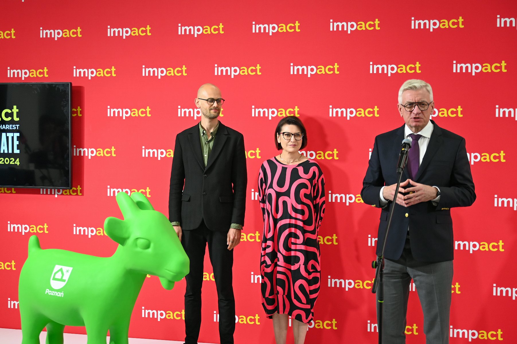 Prezydent Jacek Jaśkowiak przemawia podczas międzynarodowego kongresu "Impact'24". Obok niego stoją dwie osoby: kobieta i mężczyzna. Na dole po lewej stronie zielona figura koziołka z nowym logo. - grafika artykułu