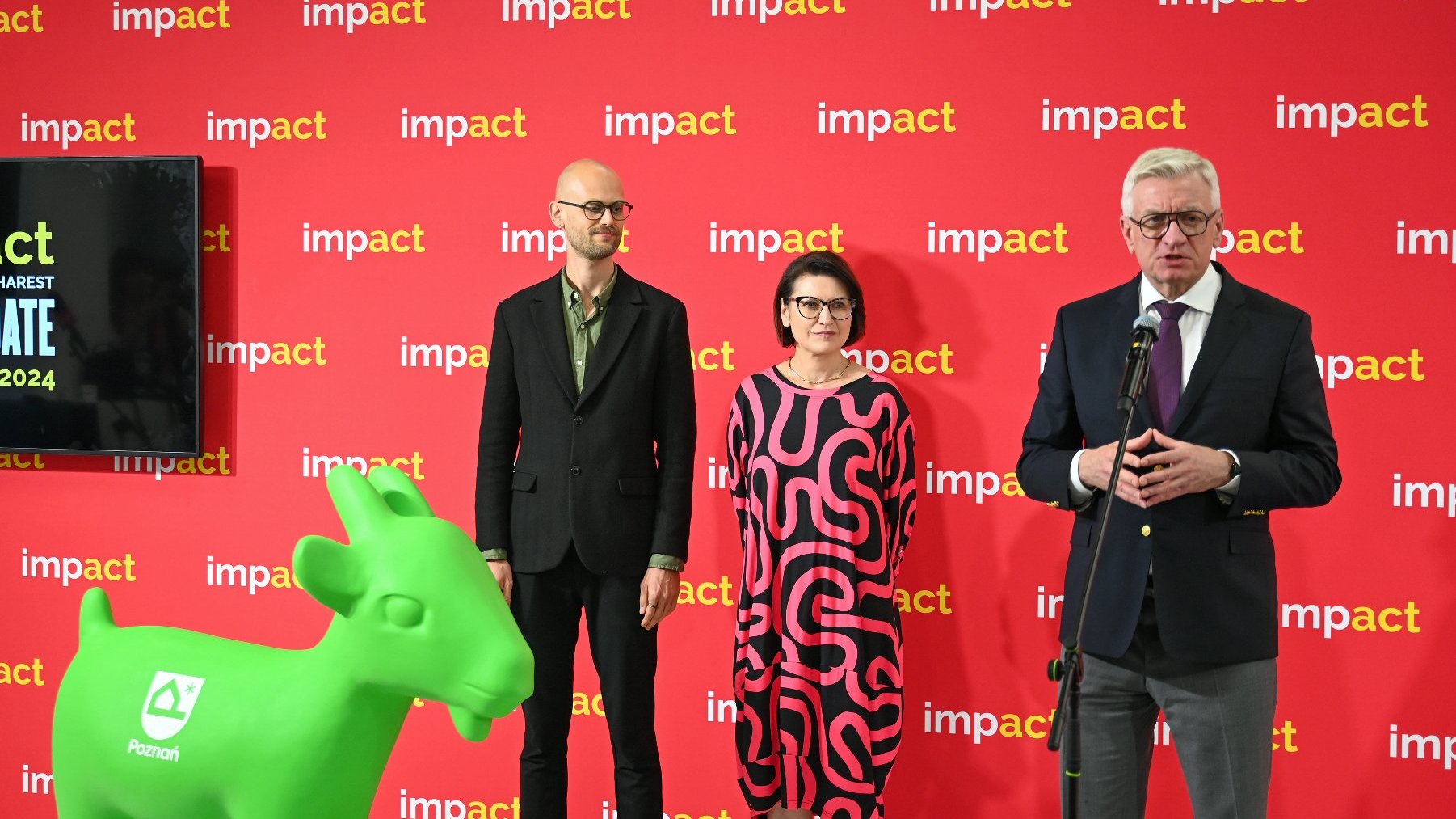 Prezydent Jacek Jaśkowiak przemawia podczas międzynarodowego kongresu "Impact'24". Obok niego stoją dwie osoby: kobieta i mężczyzna. Na dole po lewej stronie zielona figura koziołka z nowym logo.