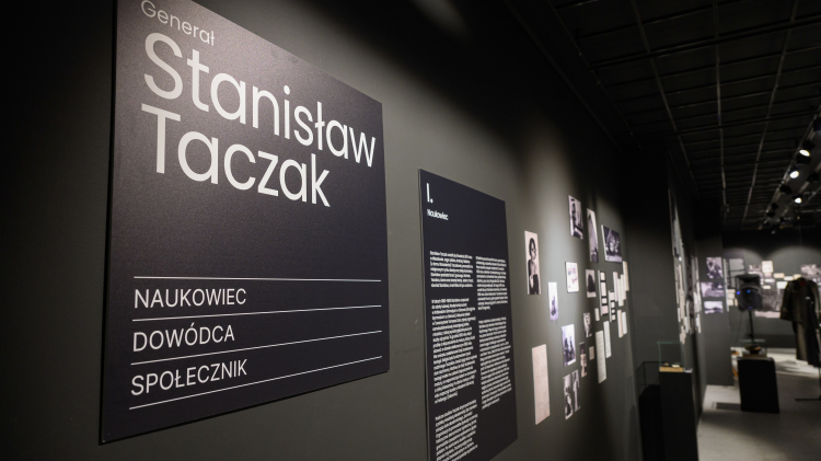 Fragment ekspozycji: czarne wnętrza Muzeum, na pierwszym planie tablica z tytułem wystawy, w tle na ścianie widoczne zdjęcia.