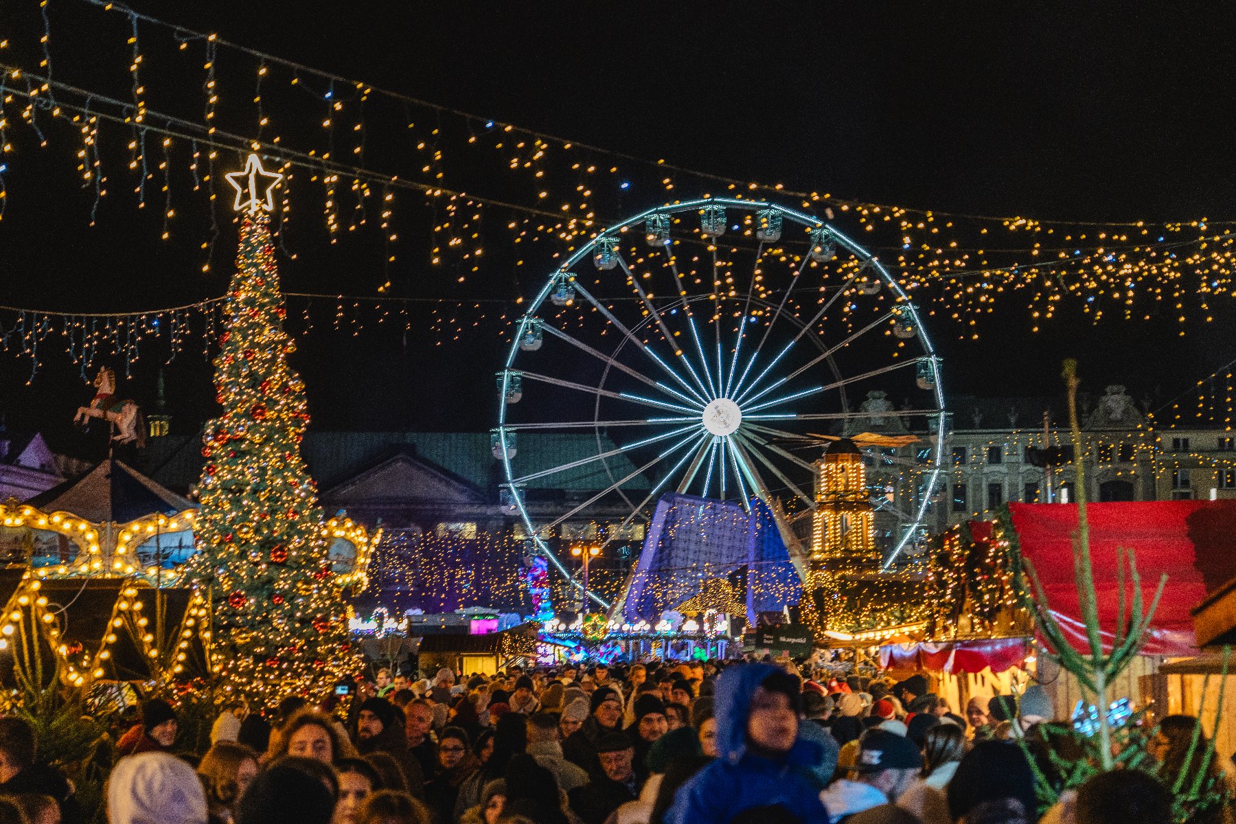 Zdjęcie przedstawia świąteczny plac Wolności. Oprócz kolorowych lampek widać na nim tłum ludzi, stoiska handlowe, choinkę i młyńskie koło. - grafika artykułu