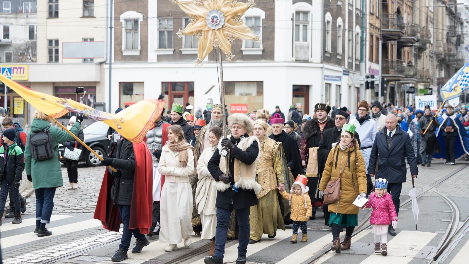 Zdjęcie przedstawia tłum ludzi idących ulicami Poznania. Osoby w pierwszych rzędach mają przebrania, niosą dużą złotą gwiazdę i żółtą flagę.