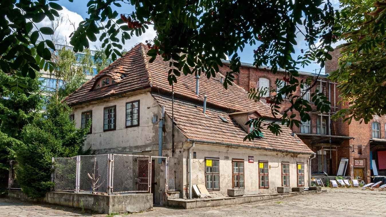 Stary, zniszczony budynek z czterema drewnianymi oknami, w tle wyższy budynek z czerwonej cegły - dawna siedziba Starej Papierni.