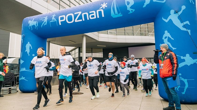 Grupa ludzi w białych koszulkach przebiega pod dużą niebieską dekoracją z logo Poznania "POZnan*".