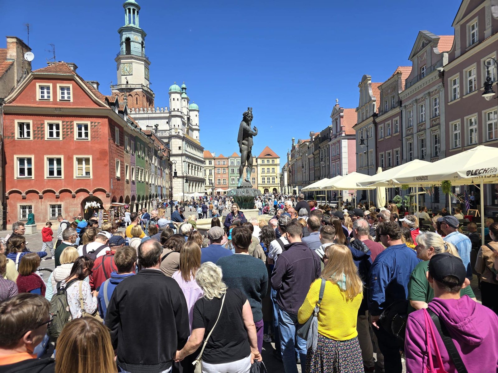 Uczestnicy wycieczki zebrani na Starym Rynku w Poznaniu. Wokół kolorowe kamienice i wieżą ratusza w tle. Na pierwszym planie znajduje się tłum zgromadzony wokół pomnika na otwartej przestrzeni, przy słonecznej pogodzie. - grafika artykułu