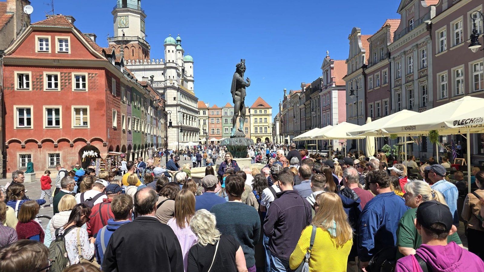 Uczestnicy wycieczki zebrani na Starym Rynku w Poznaniu. Wokół kolorowe kamienice i wieżą ratusza w tle. Na pierwszym planie znajduje się tłum zgromadzony wokół pomnika na otwartej przestrzeni, przy słonecznej pogodzie.