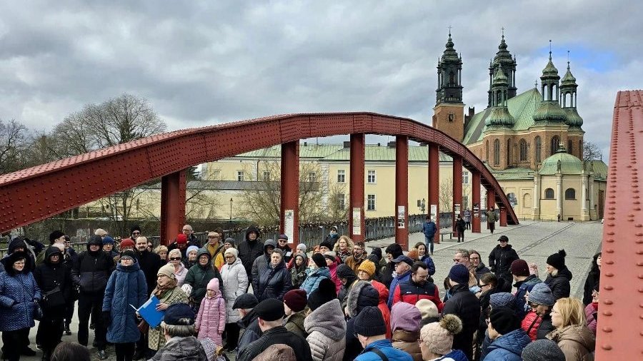 Uczestnicy przechadzki ubranych w zimowe ubrania. Zebrani na moście z czerwonymi stalowymi konstrukcjami. Przysłuchują się przewodniczce. W tle widoczna jest zabytkowa katedra z wysokimi wieżami. Na niebie dominują chmury.