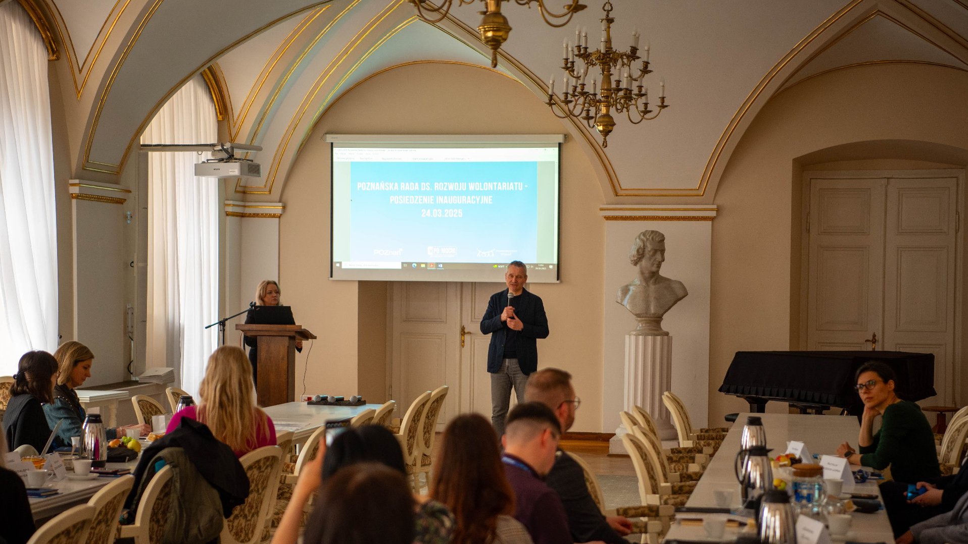 Formalne posiedzenie inauguracyjne Poznańskiej Rady ds. Rozwoju Wolontariatu. Osoby siedzą przy stole w eleganckim pomieszczeniu z łukowym sufitem, złotymi detalami oraz dużym żyrandolem. Na ekranie wyświetlony napis: ,POZNAŃSKA RADA DS. ROZWOJU WOLONTARIATU - POSIEDZENIE INAUGURACYJNE 24.03.2023'.