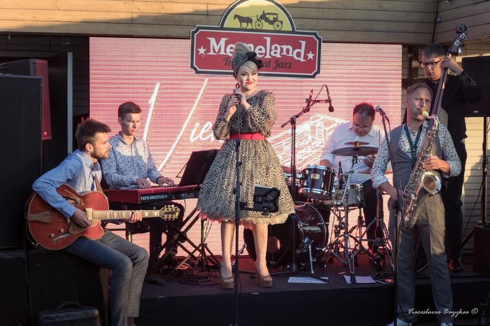 Photo of the performers on stage - a woman in a dress who is standing and holding a microphone in her hands and four men sitting and playing the musical instruments. - grafika artykułu