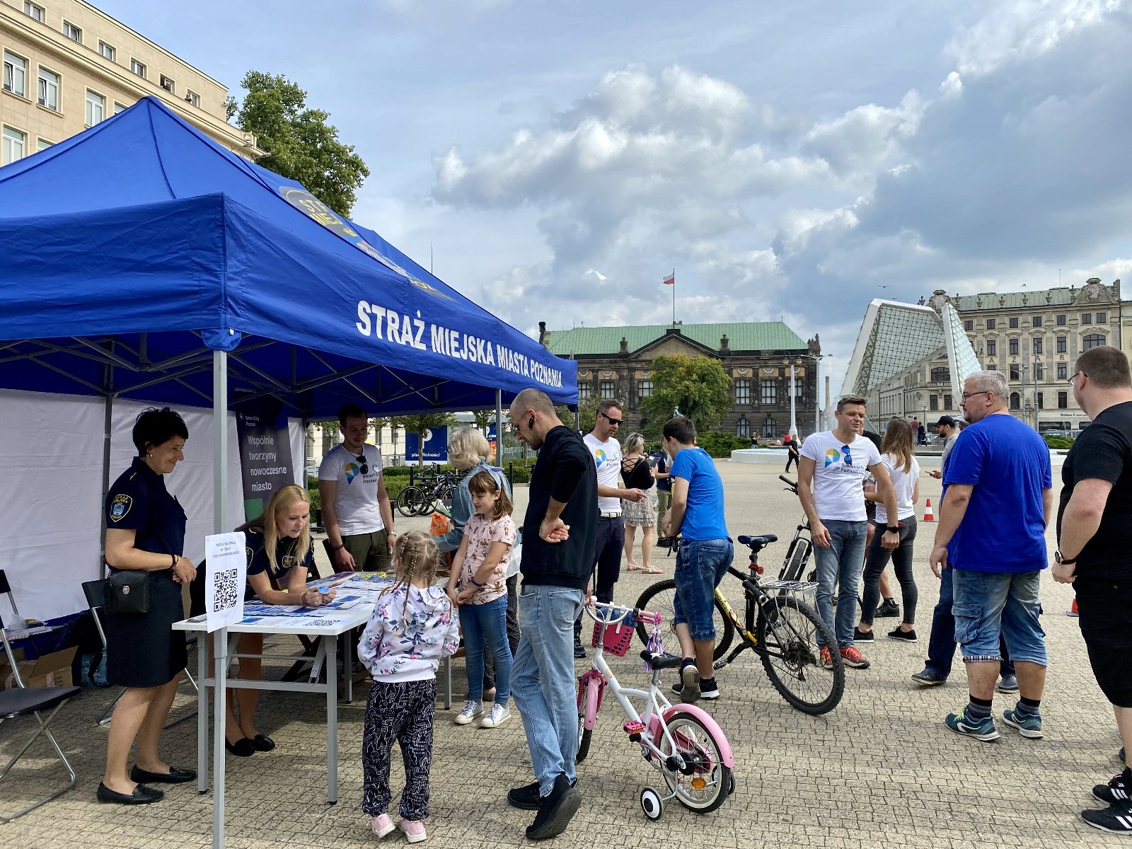 Consultation with residents, blue tent on the right, everyone standing in the square, two people holding bicycles, fountain in the background. - grafika artykułu