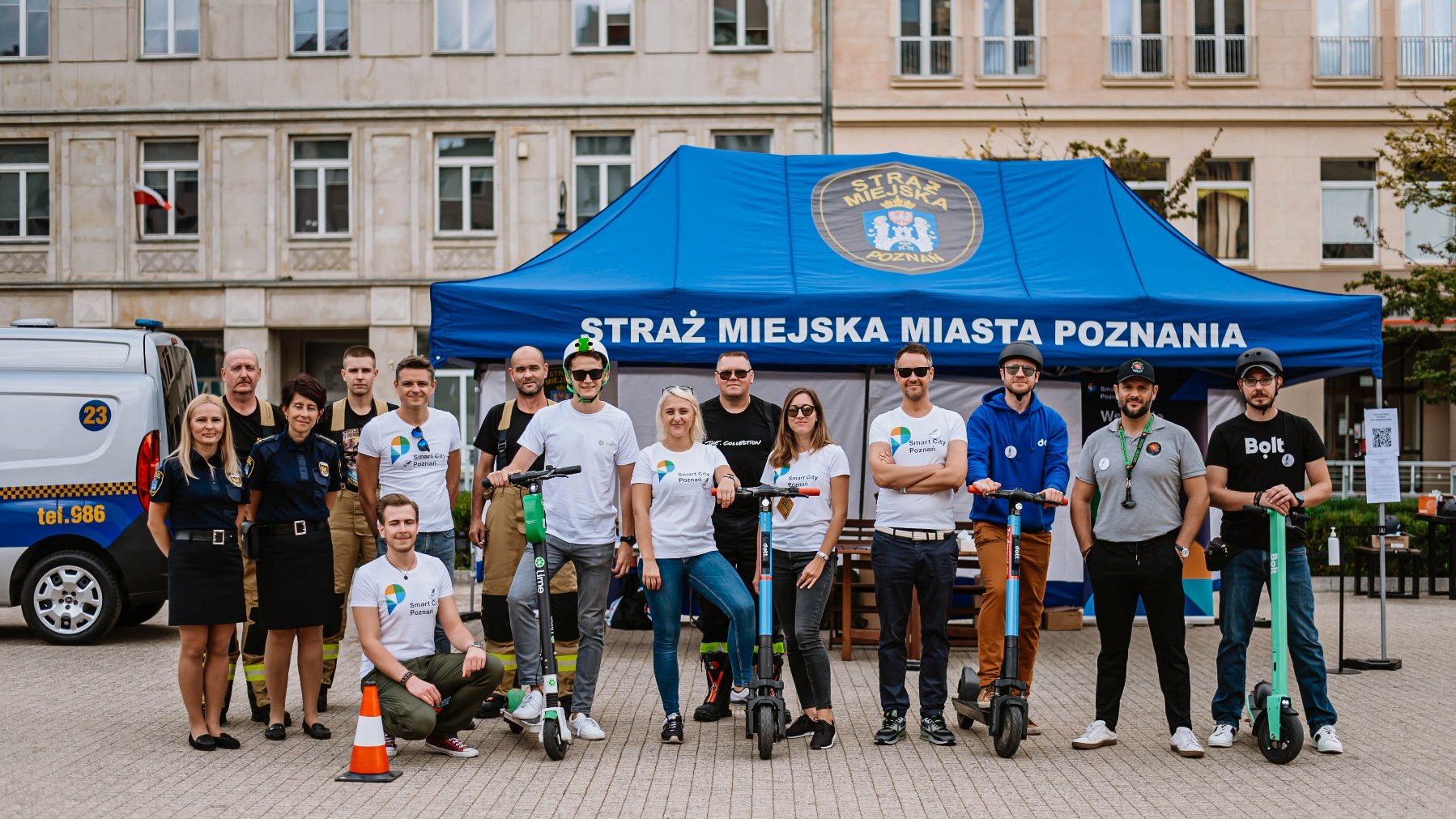 A group of people standing in front of a blue tent, some of the people are holding scooters. A building and a car in the background.