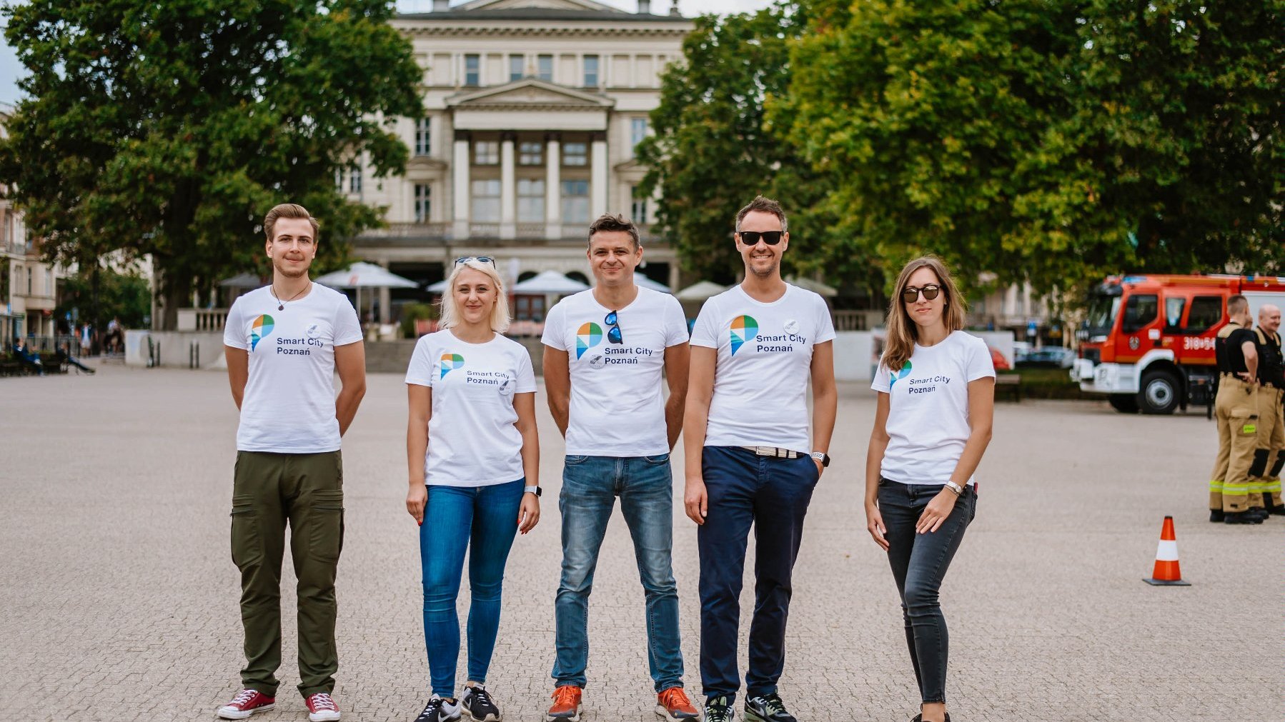 Two women and three men wearing white t-shirts with the Smart City logo, standing in the Square, with a building and trees in the background.