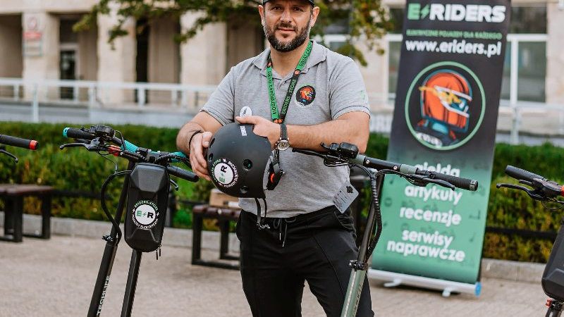 Man standing by scooters, holding helmet, wearing gray t-shirt.