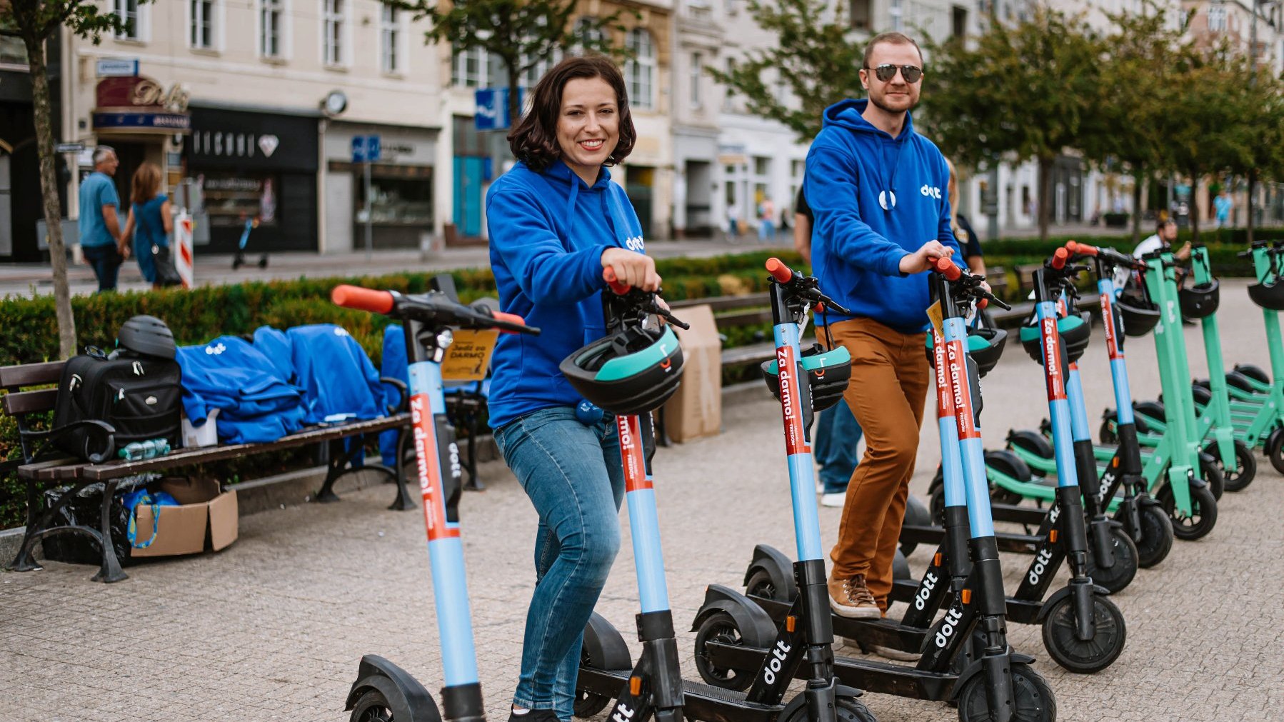 Two people in blue sweatshirts standing on scooters, with benches and other scooters in the background.