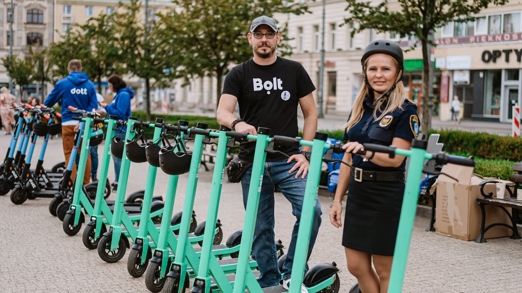 Two people by green scooters, a man in a black T-shirt and baseball cap and a woman wearing a helmet.