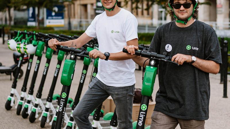 Two men wearing helmets, standing by green and white scooters.