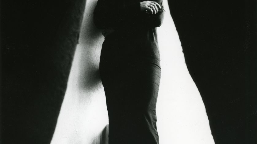 Black and white photo of Magdalena Abakanowicz - a lady dressed in black dress with her arms folded on white background. On the left and right - black curtains