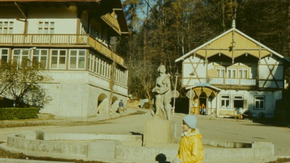 Photo of a girl sitting on a concrete parapet in front of a sculpture. Two buildings in a background.