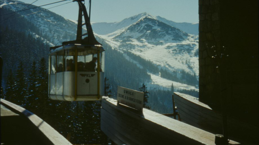 Picture of a cable car reaching a station. Snow-peaked mountains as a background.