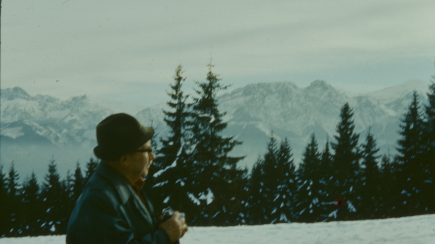 Picture of the photographer, Władysław Rut, in a profile, holding a camera in his hands. Green spruce trees and snow-peaked mountains as a background.