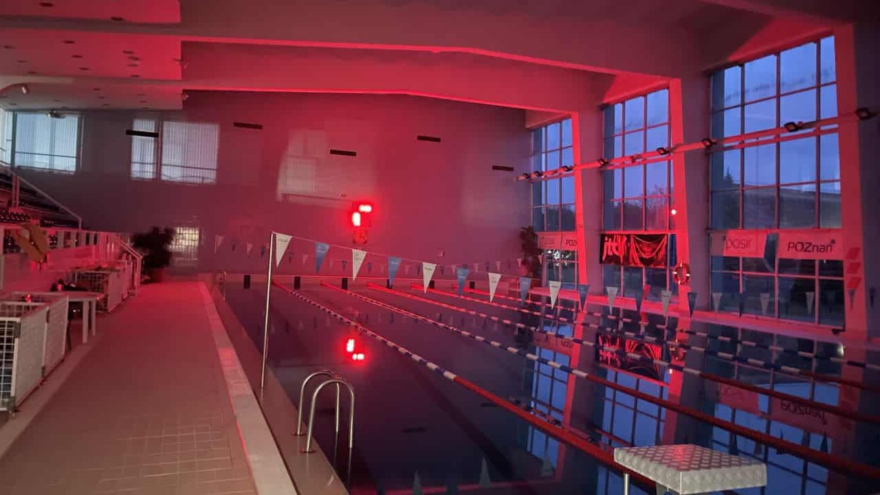 Hala basenu sportowego. Na basenie 6 torów oddzielonych kolorowymi liniami. Nad basenem rozwieszone biało-niebieska girlanda. Po lewej stronie trybuny. Po prawej stronie okna. Na dworze jest jasno. Na hali świeci się czerwone światło.