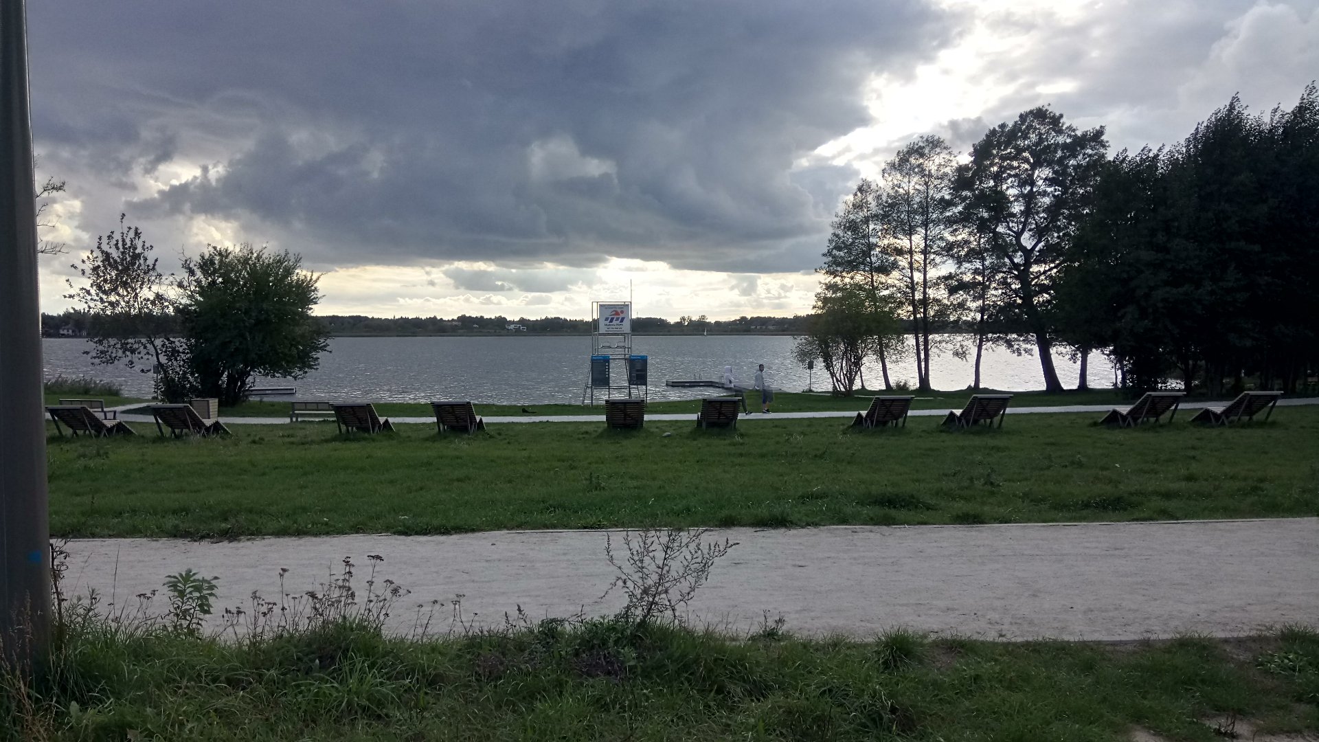 Zdjęcie przedstawia widok na Jezioro Kierskie - woda, drzewa, pochmurne niebo, leżaki