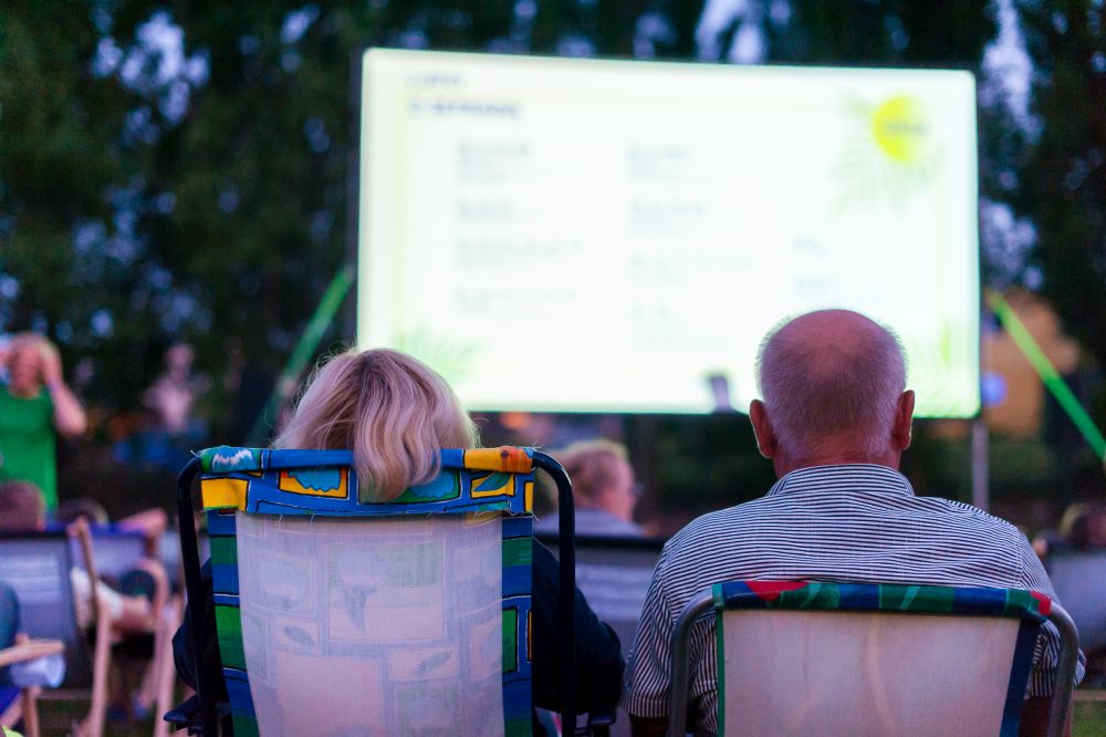 letnie kino plenerowe Na zdjęciu kobieta i mężczyzna siedzący na leżakach tyłem do obiektywu, oglądają film w plenerze