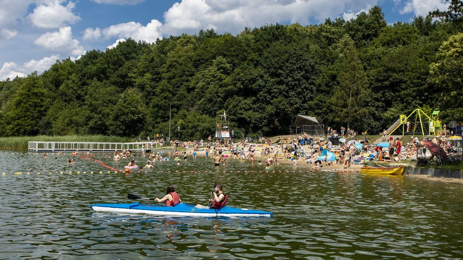 Zdjęcie przedstawia ludzi w kajaku na wodzie i tłum ludzi na plaży.